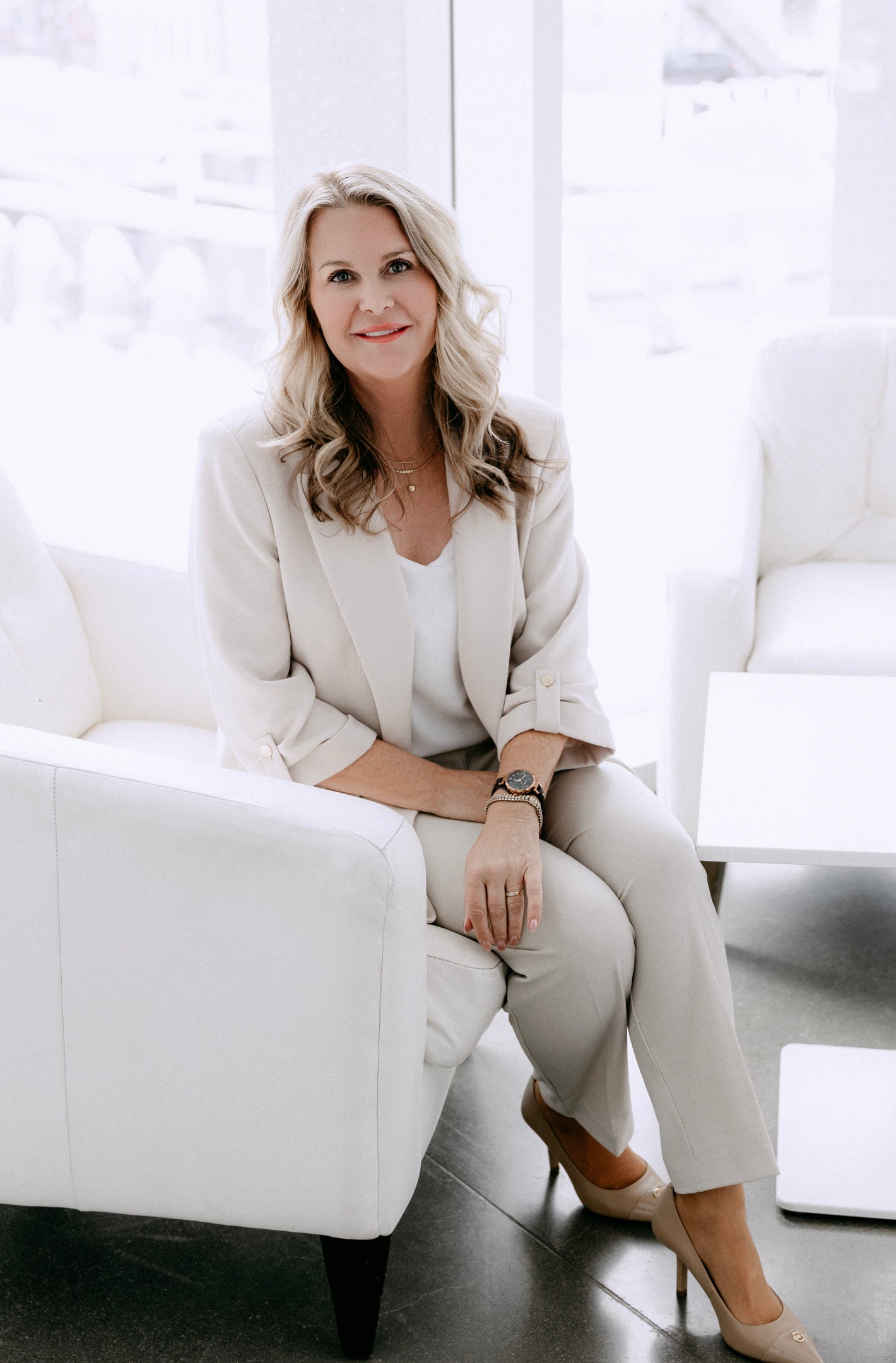 Woman in a beige suit sitting on a white couch in a bright room.